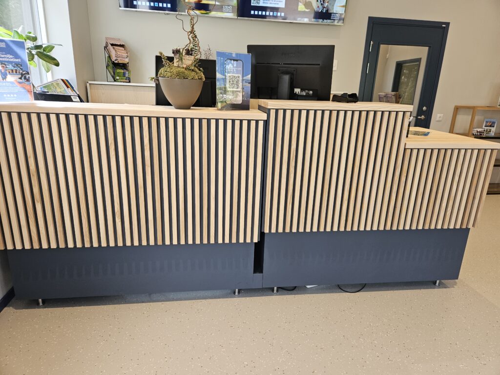 Receptionsdisk till camping med massiva askskivor och ribbor som både dämpar ljud och gör dem dekorativa. De är elektriskt höj- och sänkbara och fungerar för både sittande och stående arbete samtidigt som det uppfyller normen för tillgänglighet för rullstolsburna.